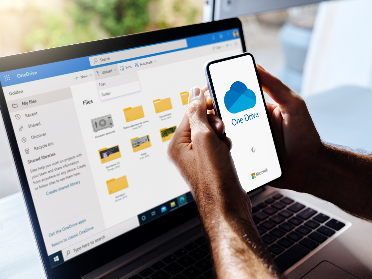 OneDrive on mobile phone and computer. Man holding a mobile phone with Microsoft cloud storage app on the screen. Laptop at background showing One Drive folfders