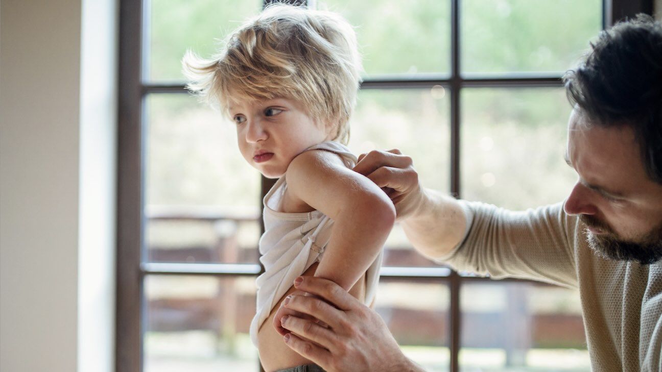 Father with small sick unhappy son indoors at home, checking his back.