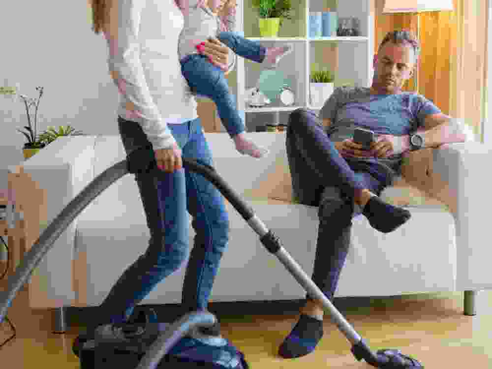 Woman with small child doing housekeeping while man sitting in couch
