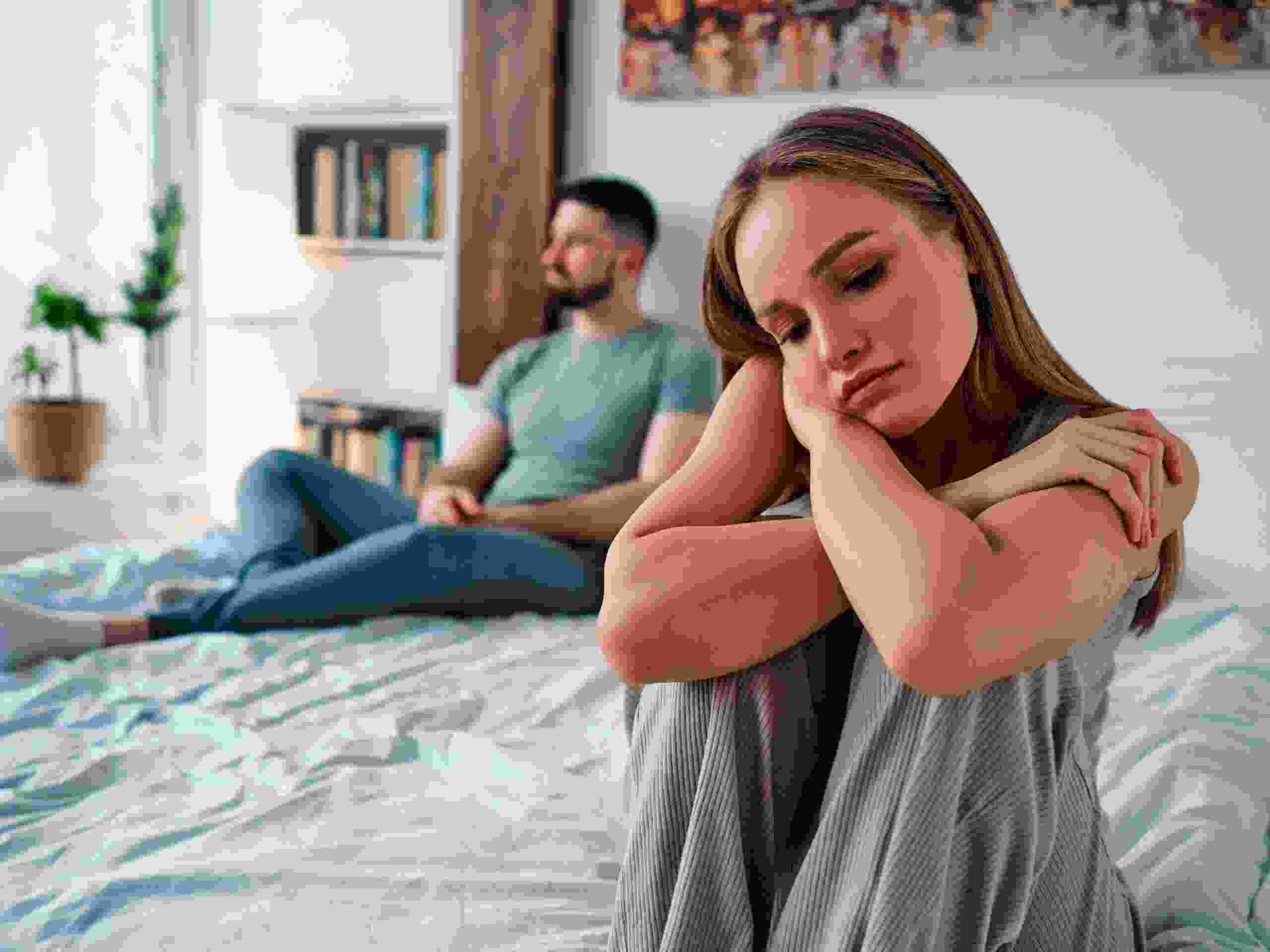 Upset young woman in home clothes sitting on the bed, in the background sitting man. Family troubles.