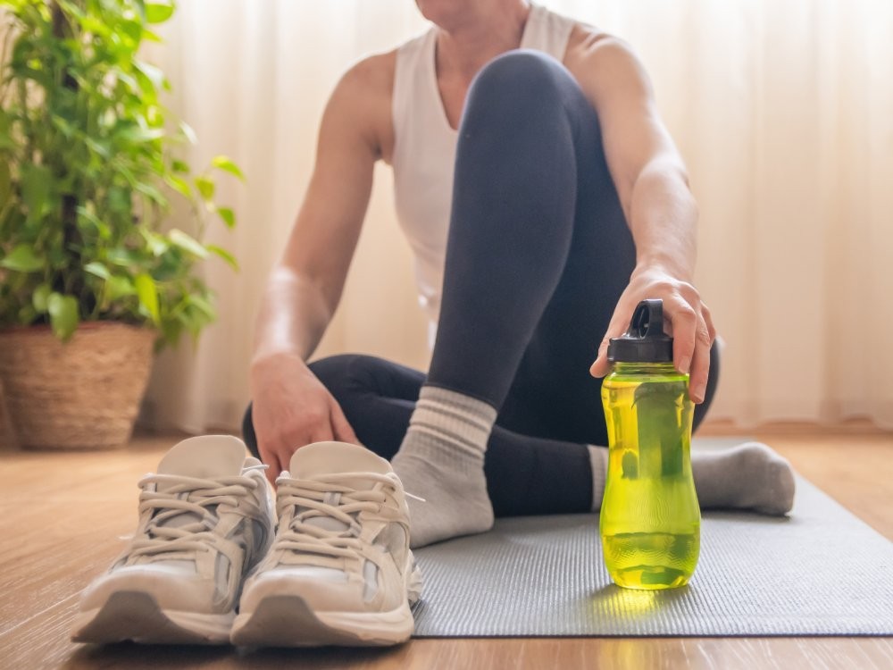 Mature woman relaxing on an exercise mat after completing a home workout, reaching for her electrolyte water bottle to stay hydrated and support her healthy lifestyle