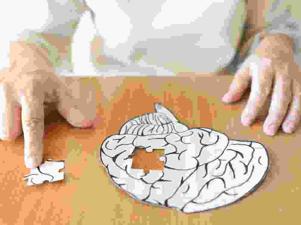 Elderly woman hands putting missing white jigsaw puzzle piece down into the place as a human brain shape. Creative idea for memory loss, dementia, Alzheimer's disease and mental health concept.